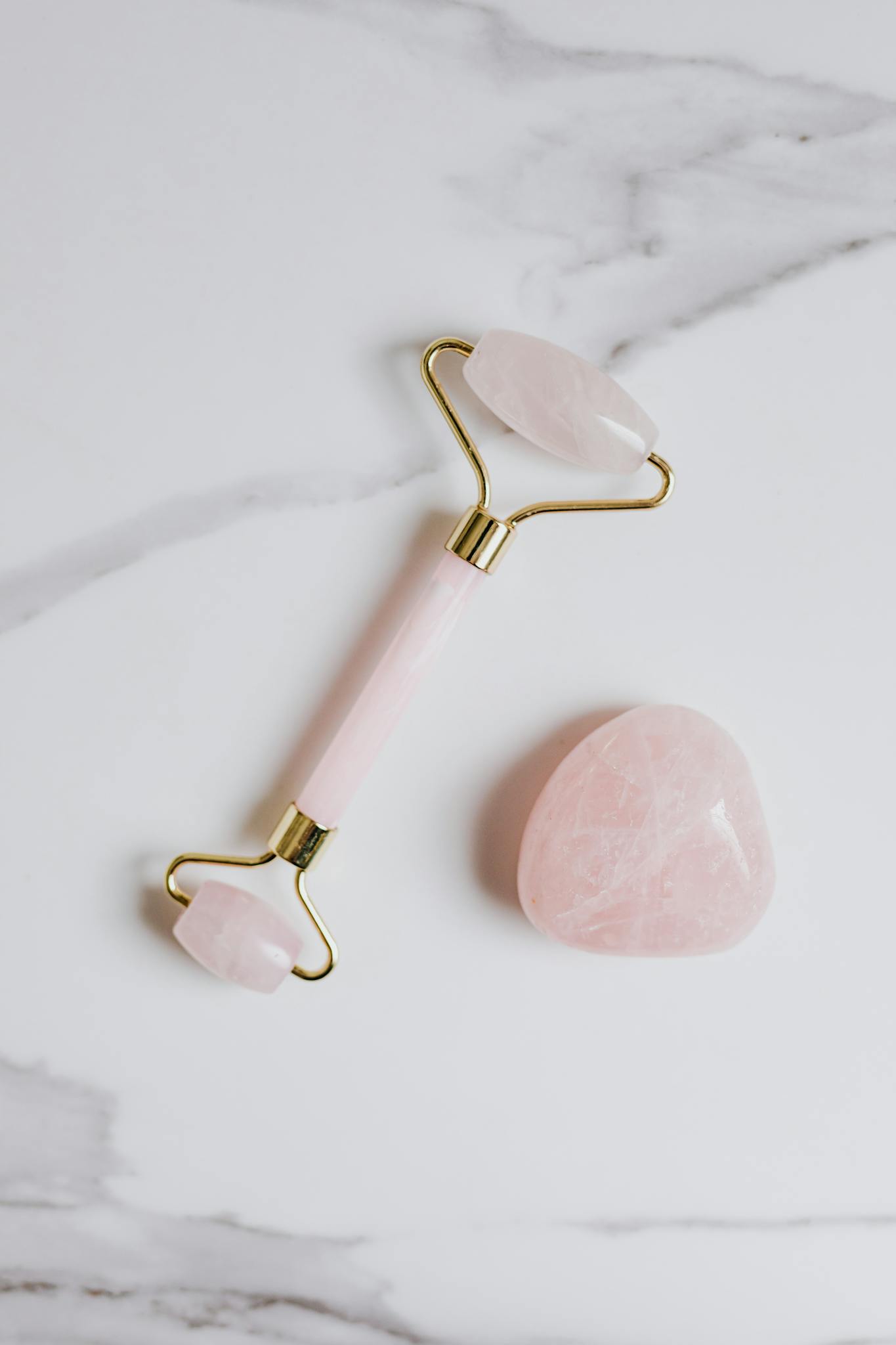 Top view of a rose quartz facial roller and stone on a marble surface.
